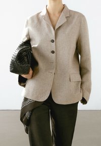 Blazer en lin marron clair à coupe ajustée, avec deux poches avant, trois boutons noirs, et une pochette tissée noire texturée. Pantalon noir avec un accent paisley.
