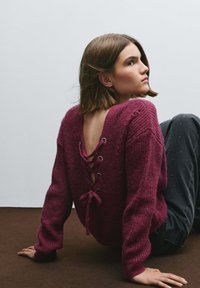 Suéter de punto color burdeos con escote en V profundo en la espalda, detalle de cordones y mangas largas. Combinado con vaqueros oscuros con acentos sutiles.