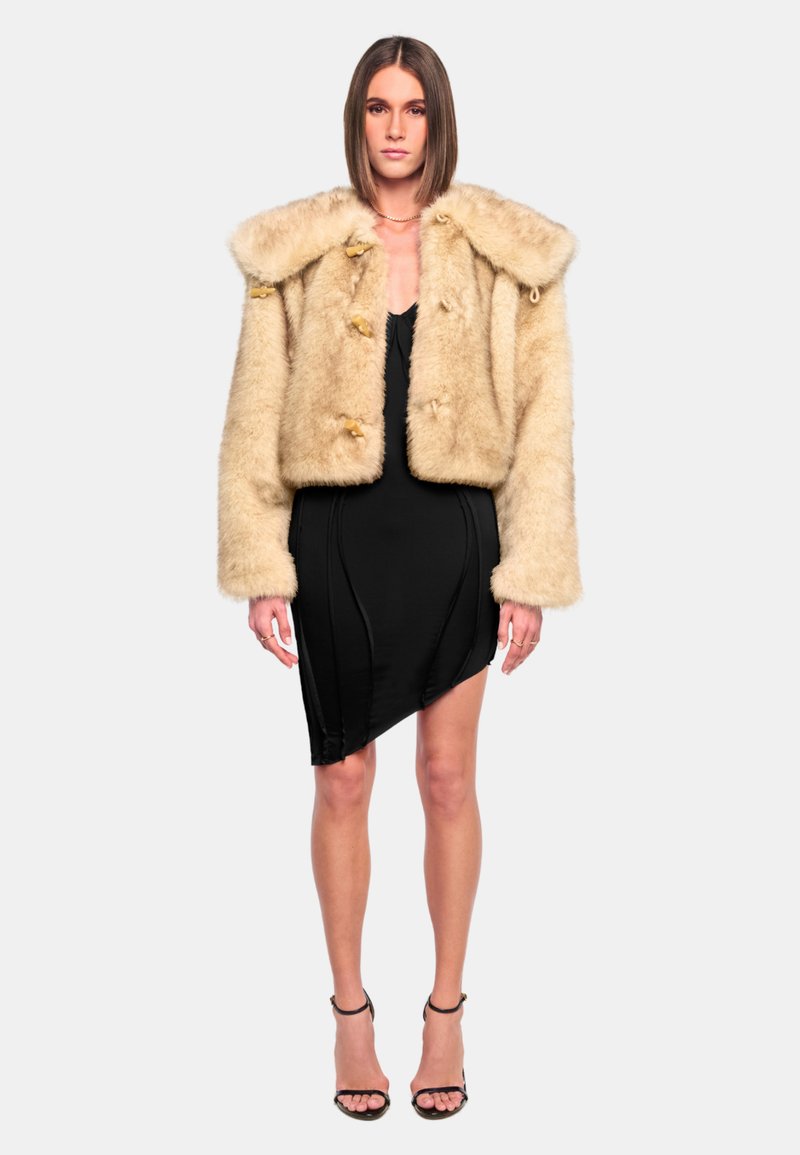 Beige faux fur cropped jacket with a large collar, button closures, and long sleeves, paired with a black asymmetrical dress and strappy heels.