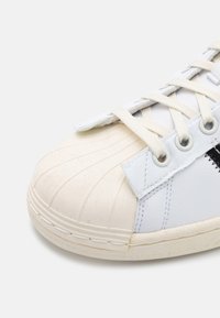 White sneaker with textured toe cap, beige rubber sole, and cream laces. Features a smooth leather body with minimal accents and stitching details.