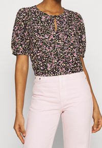 Blouse fleurie noire avec des fleurs roses et blanches, dotée de manches bouffantes et d'une encolure en forme de goutte ; associée à un jean taille haute rose clair.