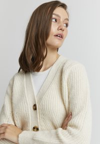 Cremefarvet strikket cardigan med V-hals, prydet med tre brune knapper og ribbet tekstur på ærmerne og kroppen.