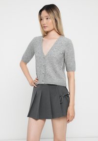 Cardigan en tricot gris à manches courtes, décolleté en V, fermeture par boutons et ornements en perles. Associé à une jupe grise plissée.