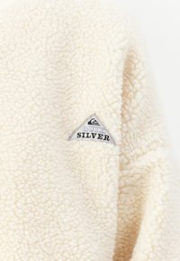 Krémová fleecová látka s malou trojuholníkovou nášivkou značky Quiksilver.