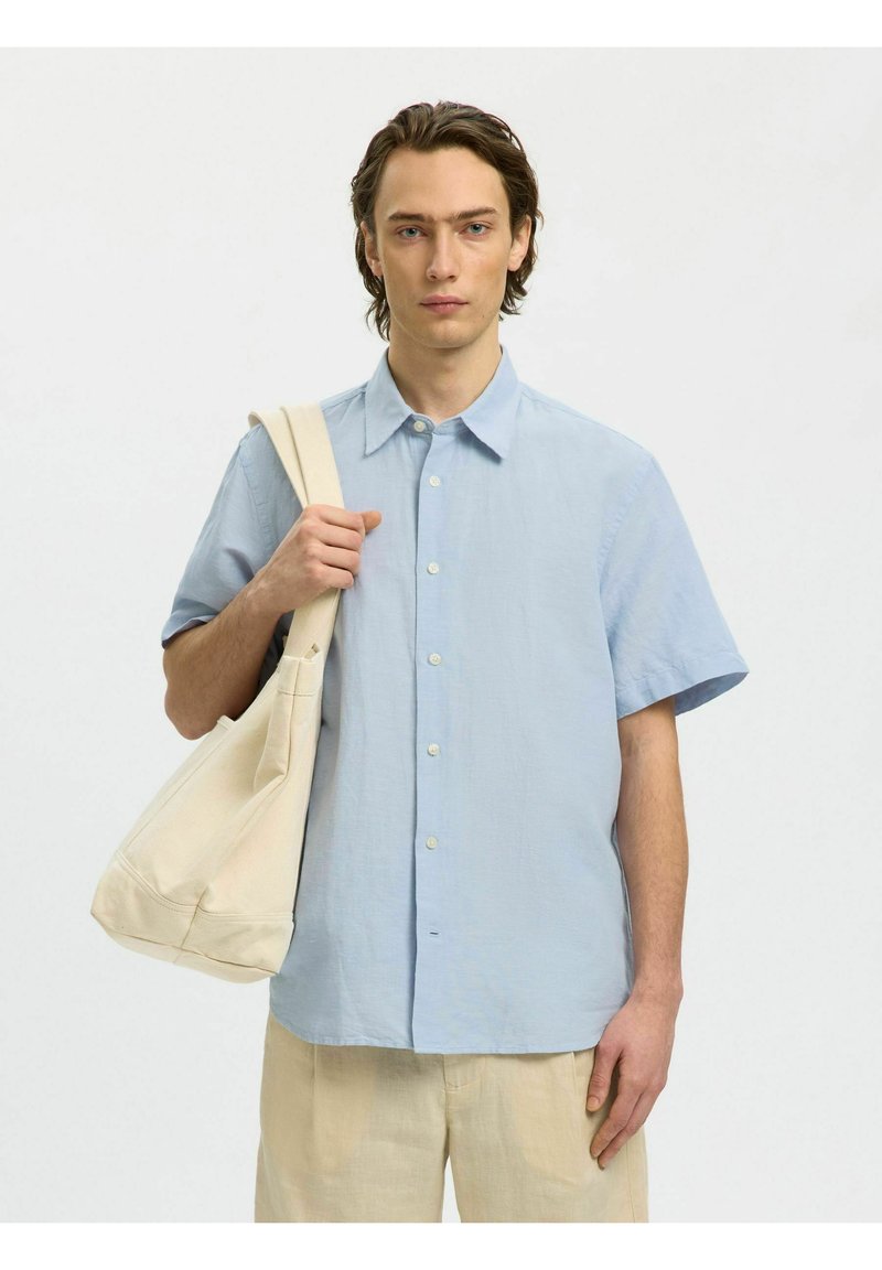 Jeune homme portant une chemise à manches courtes bleu clair et un pantalon beige, tenant un sac fourre-tout en toile crème sur l'épaule, sur un fond uni.