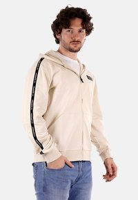 Felpa beige con zip realizzata in tessuto morbido, dotata di due tasche laterali, cappuccio con cordino e nastro di marca nero lungo le maniche.