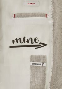 Das Innenfutter des schmal geschnittenen Kleidungsstücks ist cremefarben, mit einer gemusterten, beigen Tasche und dem aufgedruckten Text "mine" mit einem Pfeil. Rote Nähte setzen Akzente.
