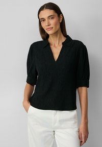 Schwarze, strukturierte Bluse mit V-Ausschnitt und kurzen Ärmeln, ausgestattet mit einem umgeschlagenen Manschettendesign, kombiniert mit weißen Hosen.