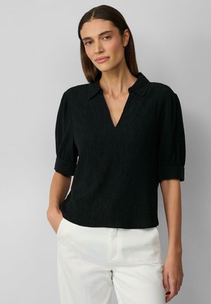 Zwarte gestructureerde blouse met een V-hals en korte mouwen, voorzien van een opgerolde manchet, gecombineerd met witte broek.