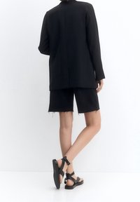 Schwarzer strukturierter Blazer, kombiniert mit schwarzen Denim-Shorts mit ausgefransten Säumen und schwarzen Riemchensandalen mit flacher Sohle.