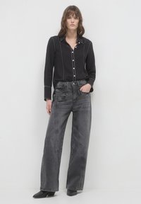Donna che indossa una camicia nera con bottoni e jeans neri a gamba larga sbiaditi, in piedi con una mano in tasca contro uno sfondo bianco. 