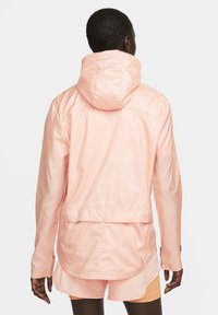 Lichte, roze, ritsjack met een capuchon, elastische manchetten en een langere achterzoom. Heeft een gestructureerd stofpatroon en een verstelbare tailleband.