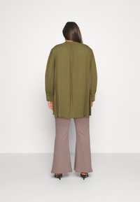 Blouse olive verte à manches longues avec des poignets boutonnés et des plis dans le dos, associée à un pantalon évasé rose clair et des talons noirs.