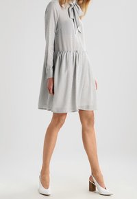 Grå randig klänning med långa ärmar, rynkad midja och rosett vid halsringningen. Bärs med vita högklackade skor.