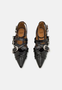Zapatos de charol negro con punta afilada, presentan tiras cruzadas y hebillas plateadas. El forro interior es marrón con el logotipo visible.