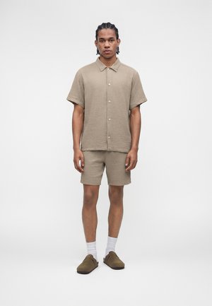 Bărbat tânăr cu părul împletit, purtând o cămașă bej cu mâneci scurte și nasturi, pantaloni scurți asortați, șosete albe și pantofi maro slip-on, stând pe fundal alb.