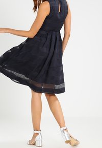 Marinblå klänning med figursytt liv och utsvängd kjol som har horisontella randiga mönster. Nyckelhål i ryggen, i kombination med silvriga klacksandaler.