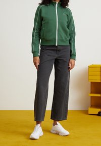 Vihreä vetoketjullinen collegepaita, jossa on valkoiset korostukset, yhdistettynä tummanharmaisiin leveälahkeisiin farkkuihin ja valkoisiin tenniskenkiin. Tasainen pinta on keltainen.