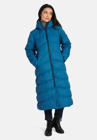 Lang, met capuchon pufferjas in een levendige blauwe kleur. Kenmerkt zich door de gequiltte textuur, volledige ritssluiting en zijzakken. Draagt over donkere broek en laarzen.