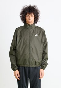 Zelenkasto-olivno Nike vetrovka s visokim ovratnikom, elastičnimi manžetami in pasom. Na prsih je bel Nike logotip. Gladka tekstura.