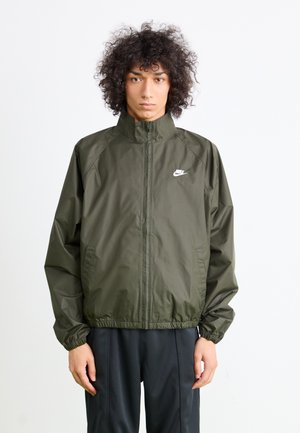 Olivengrøn Nike vindjakke med høj krave, elastiske manchetter og linning. Har et hvidt Nike-logo på brystet. Glat tekstur.
