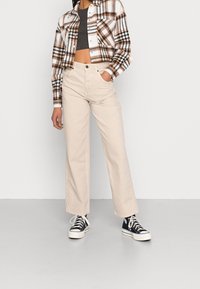 Beżowe spodnie o szerokich nogawkach zestawione z krótką, brązowo-białą kratowaną koszulą. Modelka w czarno-białych trampkach, ręce w kieszeniach.