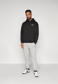 Crna dukserica s kapuljačom, s prednjim džepom i plavim tekstom "City". Uparena sa svijetlosivim treniranim hlačama i bijelim tenisicama s crnim i zlatnim akcentima.