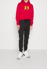 Czerwona bluza z kapturem z żółtym nadrukiem błyskawicy, czarne spodnie dresowe ze ściągaczem oraz białe trampki z czarnymi i szarymi akcentami.