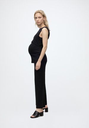 Donna incinta in piedi di profilo che indossa una canotta nera senza maniche, pantaloni neri e sandali neri con tacco su uno sfondo chiaro uniforme.
