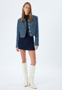 Blå tweed cropped jakke med sølvknapper, parret med en marineblå tætsiddende kjole og knæhøje hvide stiletter. Glat tekstur hele vejen igennem.