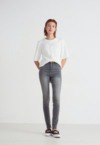 White ribbed short-sleeve cardigan with buttons, paired with slim gray jeans and black slip-on shoes. Simple, casual outfit on a light background.