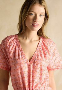 Ružový kvetinový šaty s výstrihom do V, krátkymi naberanými rukávmi a zúženým pásom. Materiál je ľahký a textúrovaný.