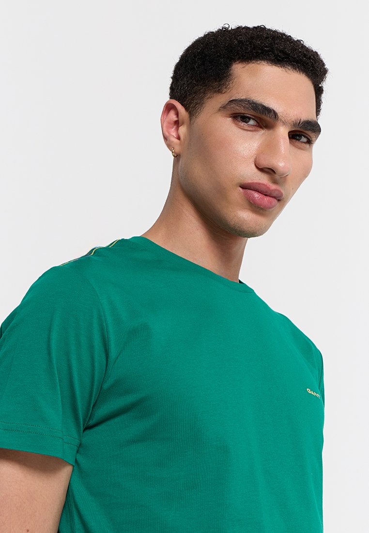 Camiseta de algodón verde con cuello redondo y mangas cortas. Presenta un detalle minimalista de logo dorado y costuras visibles en el cuello.