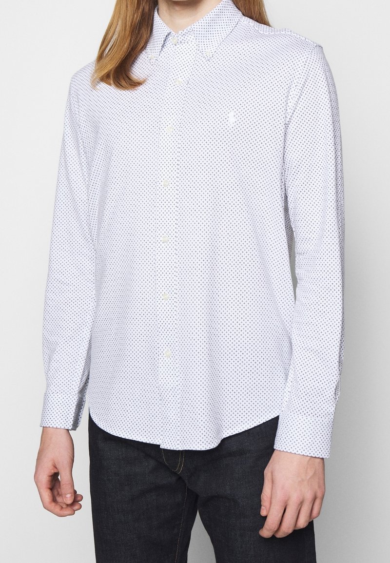 Muž ve bílé košili s dlouhým rukávem, která má malý námořnický modrý diamantový vzor, a tmavých džínách, stojící na pozadí bez vzoru.