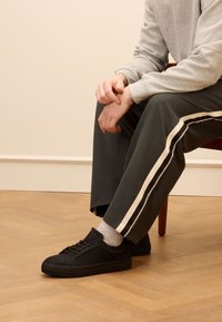 Zwarte suède sneakers met een platte zool, grijze enkelsokken, in combinatie met donkergroene broek met witte zijstrepen en een grijze sweatshirt.