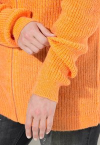 Cardigan arancione lavorato a maglia con cerniera e maniche lunghe a costine. Il tessuto è testurizzato, mostrando un colore caldo e vibrante.