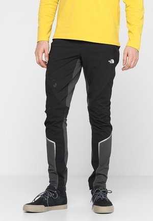 Mężczyzna ubrany w czarne i szaro-szare sportowe spodnie marki North Face, żółtą koszulkę z długim rękawem oraz czarne trampki z beżowymi podeszwami.