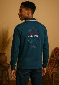Man in een donkergroene lange mouw shirt met "J&JOY" en een patroon op de achterkant, staande in een kamer met geweven decor en een gebreide deken.