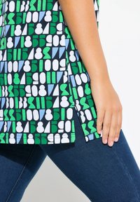 T-shirt met korte mouwen met een geometrisch patroon in groen, blauw en wit op een donkere achtergrond met zijsplitten; gecombineerd met donkere spijkerbroeken.