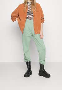 Pomarańczowa kurtka z sztruksu na zamek, wzorzysty top z siateczki, mięta zielone spodnie dresowe i czarne masywne buty o fakturowanym wykończeniu.