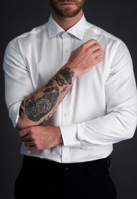 Witte lang-omslagen dressshirt, soepele stof, met knoopkraag, gecombineerd met zwarte broek, voorzien van zichtbare donkere bloemen tatoeages op de onderarm.