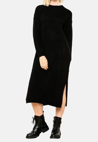 Svart stickad klänning med långa ärmar, rund halsringning och sidoklyftor. Åtsido med svarta snörboots. Mjuk textur.