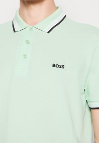 Gros plan d'une personne portant un polo vert clair à trois boutons avec "BOSS" brodé sur la poitrine.