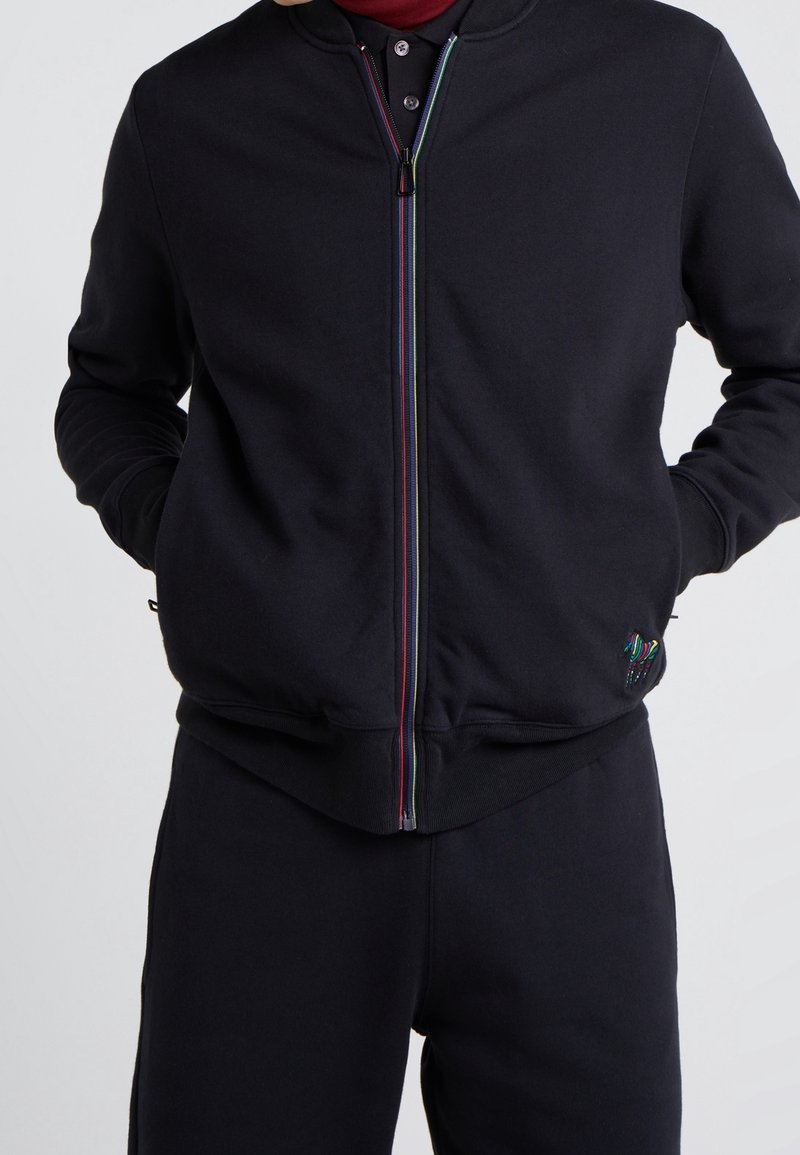 Persona che indossa una giacca nera con zip e pantaloni neri abbinati, con le mani nelle tasche della giacca, e un piccolo dettaglio ricamato colorato vicino all'orlo.