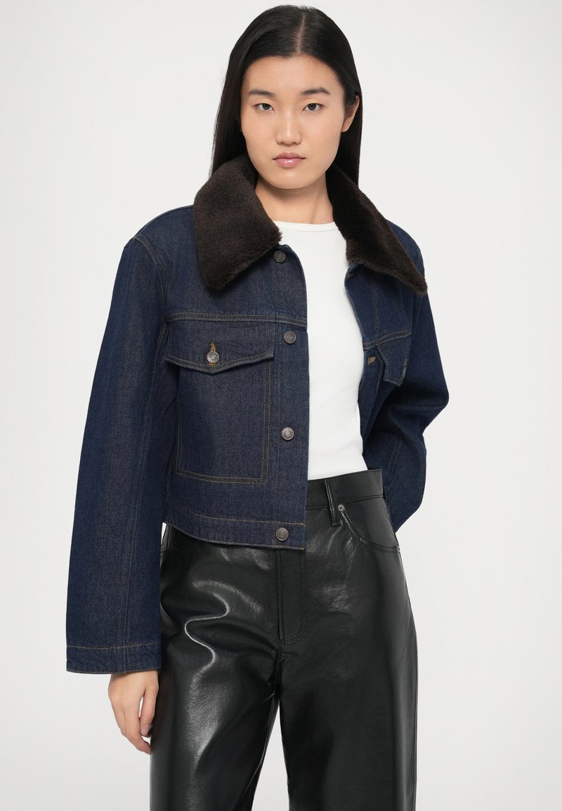 Giacca di jeans corta in blu scuro con colletto in pelliccia marrone, dotata di due tasche frontali, chiusura con bottoni e dettagli di cuciture a contrasto.