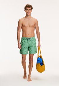 Grüne Badeshorts mit Palme-Prints, elastischem Bund und Kordelzug. Das Model hält eine gelbe Tasche mit blauer Kleidung.
