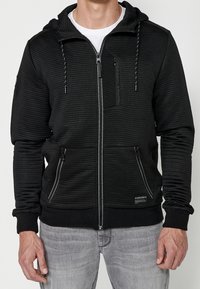 Felpa con cappuccio nera con zip, realizzata in tessuto a coste testurizzato, cappuccio regolabile con coulisse e due tasche laterali con zip. Presenta una toppa con logo.