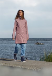 Vattentät rosa regnrock med hög krage, utrustad med blixtlås framtill och fickor, kombinerad med blå jeans och marinblå gummistövlar.