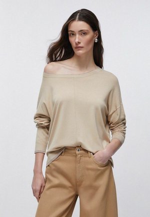 Vrouw met lang donker haar, draagend een beige off-shoulder trui en lichtbruine broek, staand met één hand in de zak tegen een effen achtergrond.