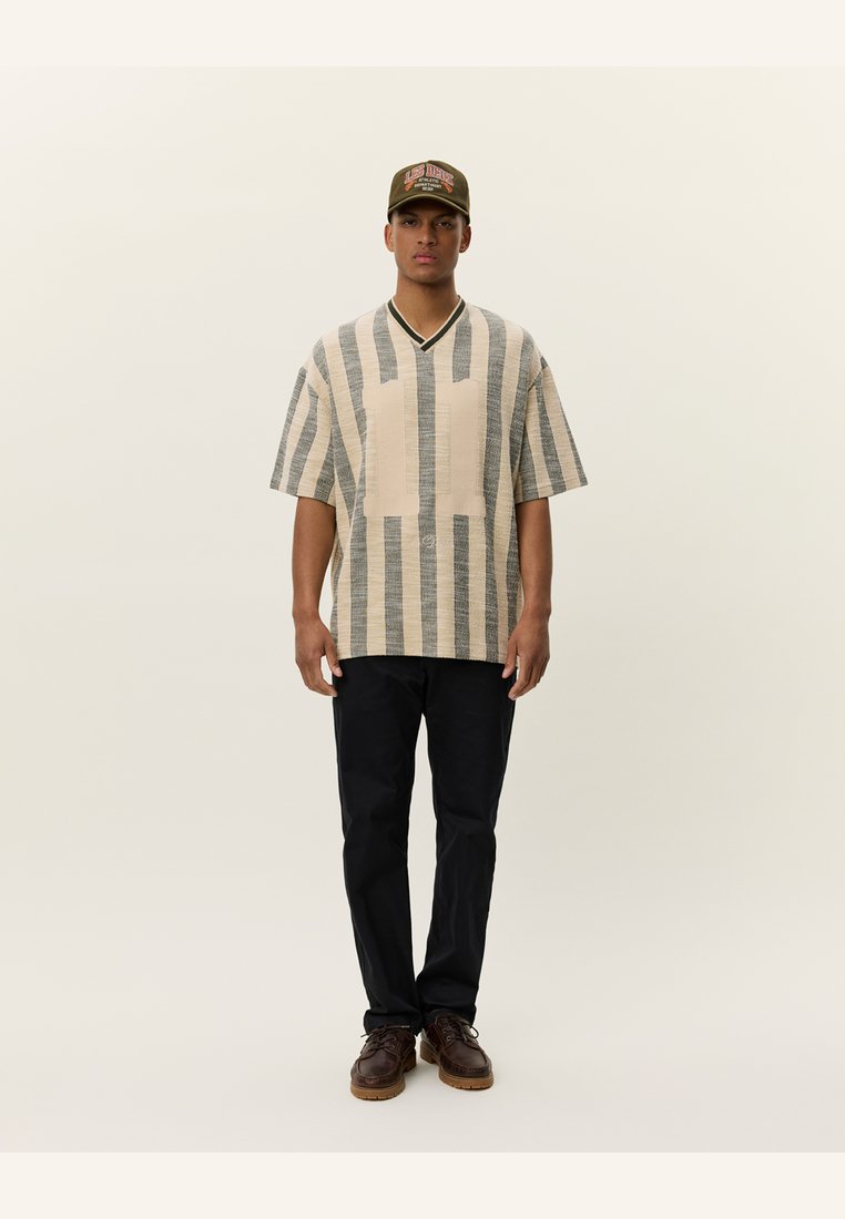 Light beige oversized shirt with navy stripes, V-neckline, featuring a single horizontal pocket. Paired with black trousers and brown shoes.